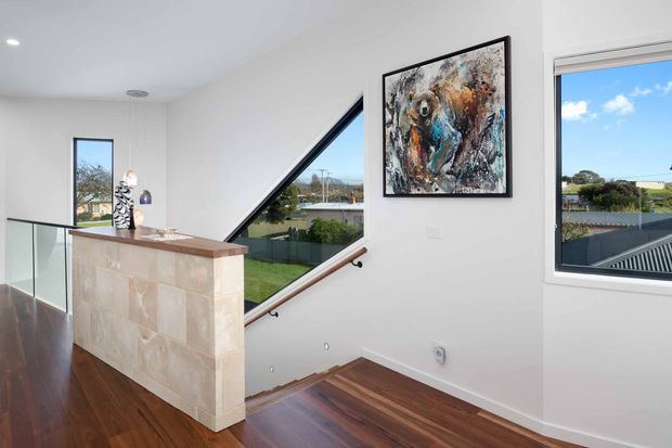 Bright contemporary stair landing with white walls, dark polished timber flooring, a glass balustrade, and a tiled half-height feature wall topped with a timber ledge. A timber handrail follows the stairs beside a large angled window, while additional windows provide views outside. Abstract artwork and small decorative objects add colour to the otherwise clean, light interior.