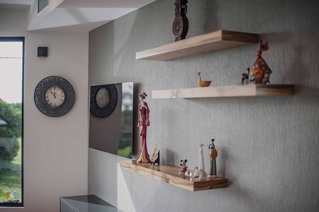 Interior vignette showing a grey textured feature wall with three floating timber display shelves holding decorative objects. A large round wall clock, mirrored panel, narrow window, and low dark console are also visible. The space is softly lit and appears to be a hallway or living-area display zone.