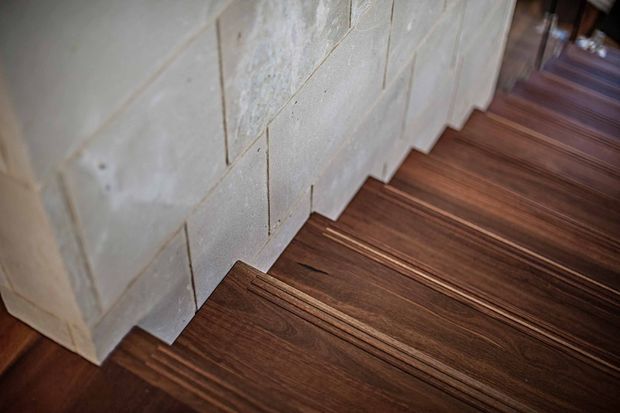 Close-up architectural detail of a staircase with dark timber stair treads and risers beside a pale, textured masonry or stone wall. The image emphasises the contrast between the warm wood grain and light wall surface; no furnishings or broader room context are visible.
