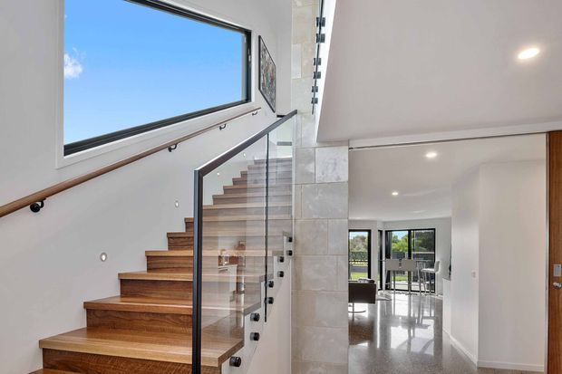 Bright contemporary interior view focused on an open timber staircase with a clear glass balustrade and dark metal fittings. A large horizontal window brings in blue-sky daylight, while a pale stone-clad structural wall divides the stair from an open living or dining area with polished flooring and extensive glazing. Recessed ceiling lighting and clean white walls create an airy, refined atmosphere.