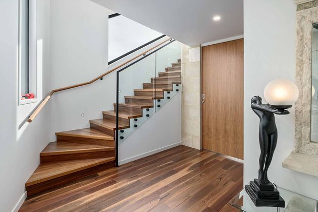 A refined residential stairway with open-riser timber stairs, matching timber flooring, and a clear glass balustrade supported by dark fittings. A timber handrail follows the stair and wall, while a plain white ceiling and walls create a bright backdrop. A timber pivot-style door and pale stone-clad wall edge are visible, along with a sculptural black floor lamp.
