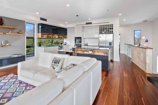 Bright open-plan contemporary interior showing a living room in the foreground flowing into a modern kitchen and dining area. White sectional seating, colourful patterned rug, timber flooring, grey walls, open shelving, dark kitchen island cabinetry, white cabinetry, stainless-steel appliances, and a timber-topped island are visible. A glazed stair balustrade and hallway appear to the right.