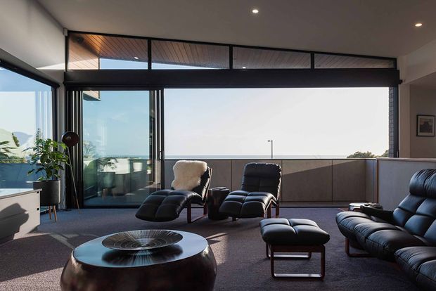 A sophisticated living room with expansive floor-to-ceiling glazing and an open ocean-facing outlook. The room features dark leather lounge chairs and sectional seating, a large round coffee table, charcoal carpet, dark window frames, and a low balcony or parapet beyond the glazing. Warm timber-lined ceiling panels appear above the window band, while recessed ceiling lights provide subtle illumination.
