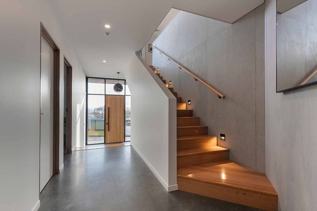 Modern residential entry hall with a sculptural timber staircase rising along a smooth grey wall. The stair treads feature warm integrated lighting, with a timber handrail and dark framed glazing at the front entrance. A polished grey floor, white walls, recessed ceiling lights, and minimalist detailing create a clean, contemporary interior.
