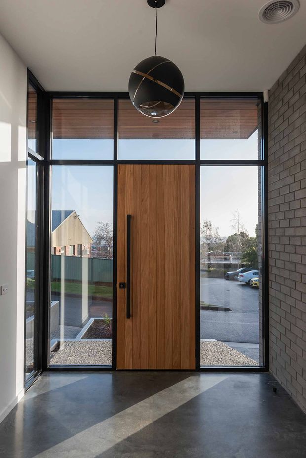 Modern residential entryway with a tall timber front door framed by extensive black-framed glazing. The entry has a polished dark concrete floor, a dark grey brick feature wall on the right, white plaster walls, and a black spherical pendant light. Exterior paving, neighbouring houses, and parked cars are visible through the glazing.