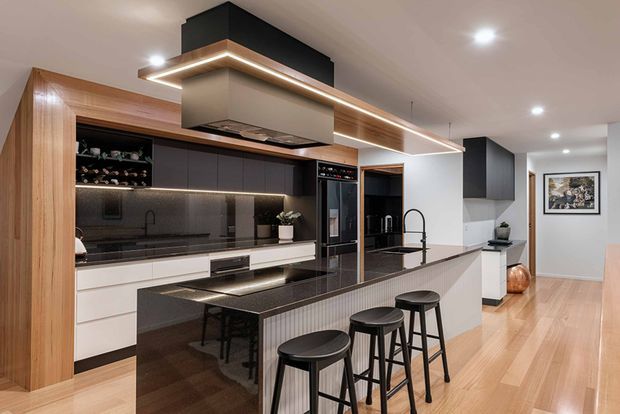 Contemporary open-plan kitchen with a large dark stone island and seating for four, white and charcoal cabinetry, integrated appliances, warm timber wall panelling and flooring, and a dramatic suspended rangehood with linear LED lighting. The kitchen opens toward a hallway and adjacent living area, creating a refined, high-contrast interior.