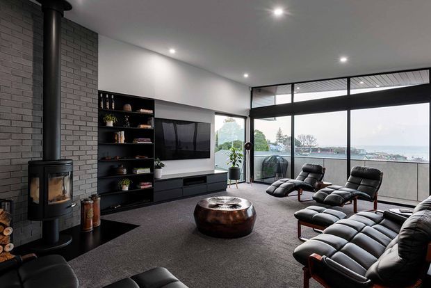 A contemporary living room with a dark grey brick feature wall, freestanding wood-burning fireplace, black built-in shelving and media cabinetry, and expansive black-framed glazing overlooking a body of water. The room is furnished with black leather lounge chairs and ottomans around a low round coffee table, with recessed ceiling lighting and a dark carpet creating a refined, moody atmosphere.