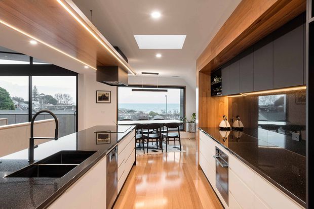 Contemporary open-plan kitchen with a long central island, black stone benchtops, integrated sink and tap, white lower cabinetry, dark upper cabinetry, and extensive warm timber joinery with concealed LED lighting. The polished timber floor leads to a dining area and large glazed doors with a water view beyond. A skylight and recessed ceiling lights provide additional illumination.