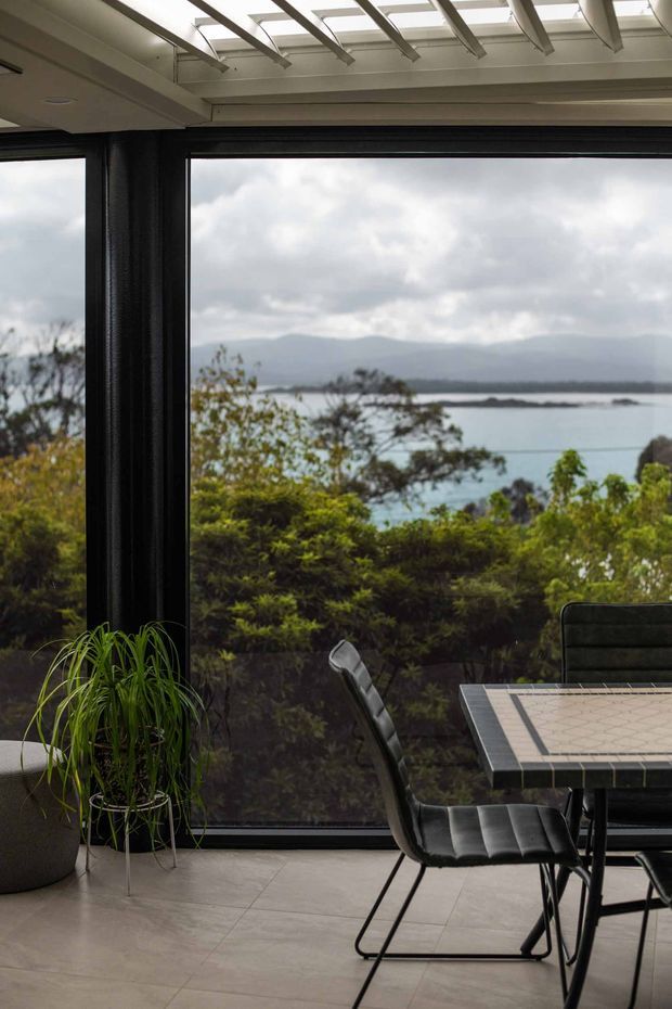A contemporary dining area beside expansive black-framed glazing, overlooking a calm coastal or lakeside landscape with mountains beyond. The room features a tiled floor, a dark tiled dining table with upholstered black dining chairs, indoor planting, and a translucent roof structure with exposed linear beams that brings in natural light.