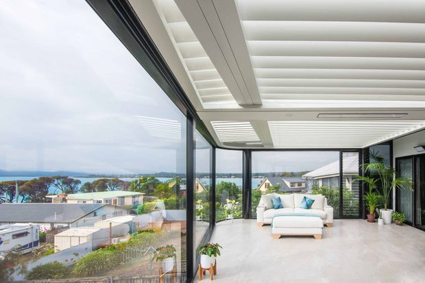 Bright enclosed outdoor living area with full-height glazed walls overlooking a coastal suburban landscape. The space features a white outdoor sofa with turquoise cushions, potted plants, pale flooring, and a distinctive white louvred pergola-style ceiling with integrated lighting and ventilation. Black-framed glazing creates a seamless connection to the view.