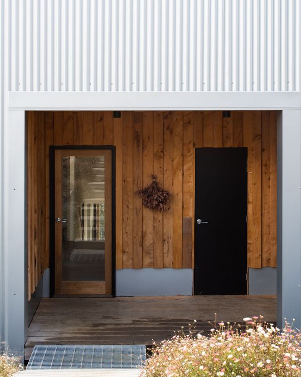 A sheltered residential entry porch framed by pale blue-grey vertical corrugated cladding and structural trim. The recessed entry has a dark timber plank feature wall, a glazed timber-framed door, and a solid black door, opening onto a weathered timber deck with a metal drainage grate and flowering planting in the foreground. The composition is simple, tactile, and contemporary with strong natural timber contrast.