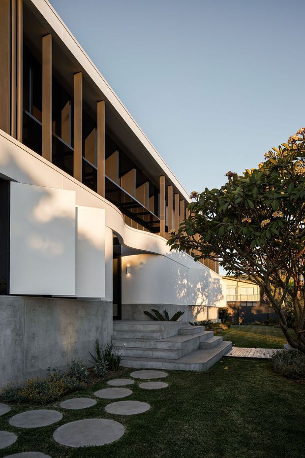 Exterior view of a contemporary two-storey building with a clean white rendered facade, exposed concrete base, long dark-framed upper-level glazing, vertical timber fins, and a curved projecting wall. Concrete steps lead through a landscaped lawn with circular stepping stones and mature planting.