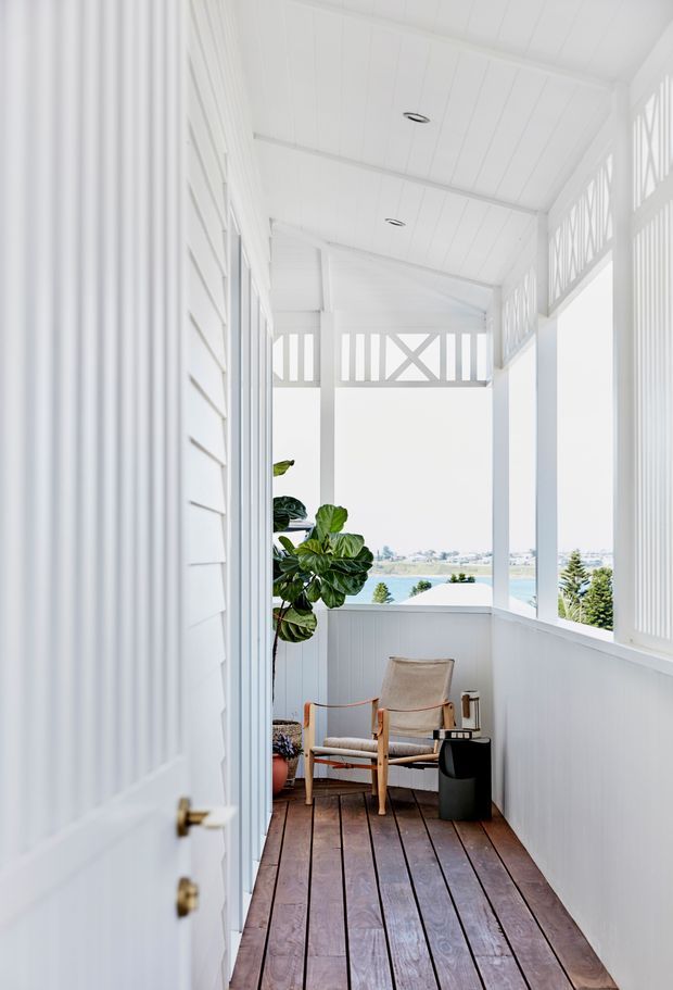 Bright enclosed outdoor verandah or balcony with white-painted weatherboard walls, decorative lattice-style upper balustrades, and a white slatted ceiling. The space has a dark timber plank floor, a timber-framed upholstered armchair, a small black side table, and potted greenery. Open sides provide a view toward water and distant buildings, creating an airy coastal setting.