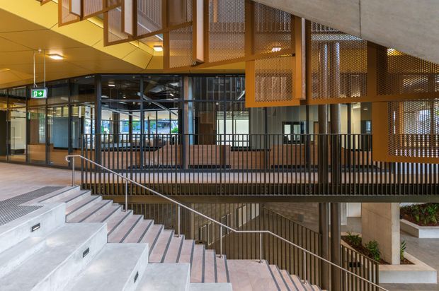 Interior view of a contemporary commercial building featuring a broad concrete stairway, black metal balustrades, timber-toned tiled flooring, perforated metal screening and suspended architectural elements. Glazed partitions reveal an open workspace or circulation area beyond, while integrated planting and strong geometric lines add visual interest.