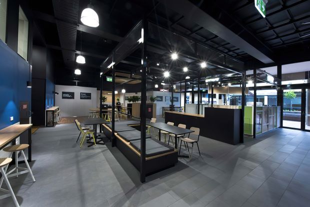 A contemporary commercial interior with an open-plan café or hospitality-style layout. The space features dark exposed ceiling services, black-framed glazed partitions, charcoal and blue walls, grey tiled flooring, communal tables, stools, bench seating, bright pendant and spot lighting, and a glazed entrance at the right.
