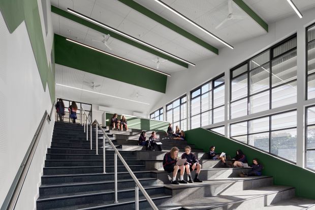 A bright, modern school interior featuring a broad stepped stairway used as informal tiered seating. Students are gathered on the dark grey steps and landings, with stainless-steel handrails, large black-framed windows, white acoustic ceiling panels, linear strip lighting, ceiling fans, and bold dark-green wall and ceiling accents.