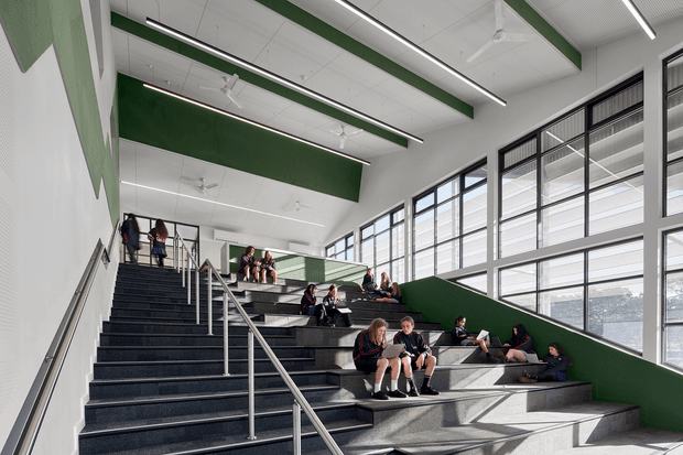 A spacious contemporary school or institutional interior featuring a broad stepped seating stairway with dark textured treads, stainless steel handrails, large black-framed windows, and green accent walls and ceiling beams. Students are seated and gathering on the tiered steps, while strong daylight enters through the glazing. The space has a bright, open, functional and communal character.