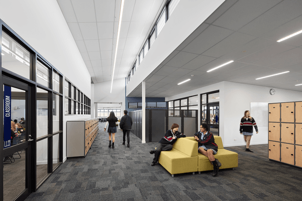 A bright contemporary school interior showing a wide carpeted circulation corridor and informal student breakout seating. Black-framed glazed classroom partitions line the left side, with timber lockers and storage along the corridor and right wall. Students occupy a vivid yellow modular lounge, while linear recessed lighting and a high clerestory-like glazed band create an airy, institutional learning environment.