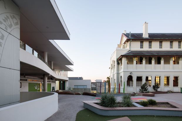 Exterior view of a contemporary multi-storey building beside a restored traditional two-storey heritage-style building. The paved courtyard includes curved raised planter beds, ornamental grasses, and colourful paving accents. The modern building has projecting horizontal floor plates, curved balconies, a pale tiled facade, and broad overhangs, while the adjacent building features cream rendered walls, a pitched roof, verandah columns, arched openings, and dark trim.
