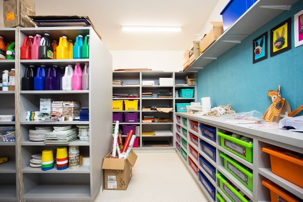 A bright, compact craft and art storage room with open shelving lining both sides and the rear wall. Shelves are filled with colourful plastic containers, paper, stationery, craft supplies, and labelled storage bins. A turquoise accent wall on the right displays framed artwork, while a long white work counter with drawers runs beneath it. The room has a practical, highly organised studio-like layout and fluorescent ceiling lighting.