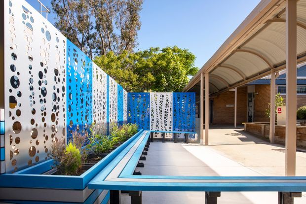 Bright exterior courtyard or covered walkway beside a brick building, featuring vivid blue raised planter beds and benches, patterned perforated metal privacy screens in white and blue, and a curved translucent canopy. Landscaping and mature trees add greenery, while the open paved area creates a contemporary civic or educational outdoor space.