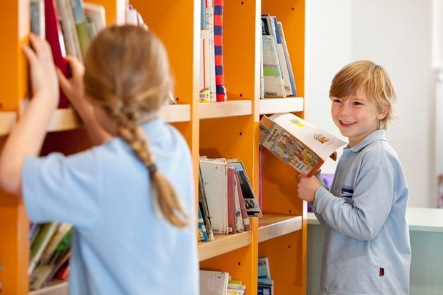 Bright photograph of two children in a colourful school or library setting, browsing books on orange built-in shelving. One child is seen from behind reaching for books, while another holds an open book and smiles. The space has vibrant orange shelving, light timber shelves, white walls, and abundant natural light.