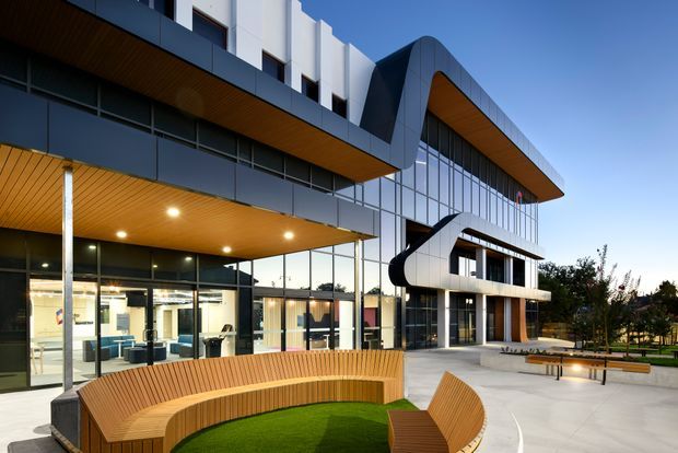 Contemporary multi-storey commercial building viewed from an landscaped outdoor courtyard. The facade combines extensive floor-to-ceiling glazing, dark metal cladding, crisp white projecting volumes, and warm timber soffits. Curved built-in timber seating surrounds a small lawn, with additional benches and paved open space in the foreground.