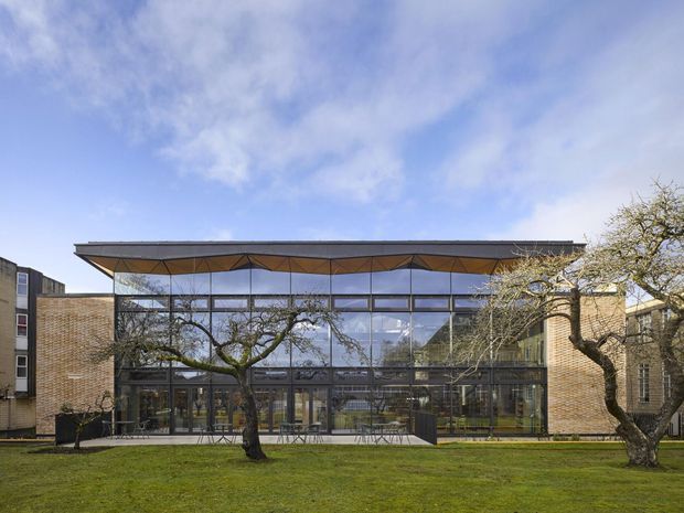 Exterior view of a contemporary institutional or commercial building with a broad flat roof, exposed angular canopy structure, extensive floor-to-ceiling curtain glazing, and warm tan brick side wings. The building fronts a manicured lawn with mature pruned trees under a bright partly cloudy sky.