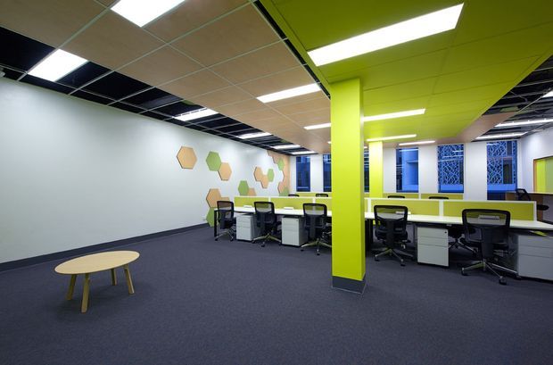 A bright open-plan commercial office with rows of white workstations and black office chairs arranged around lime-green partitions and columns. The room has dark carpet flooring, a suspended acoustic ceiling with recessed fluorescent lighting, white walls decorated with geometric hexagonal motifs, and blue-patterned windows or screens along the far wall. A small round timber table sits in the open circulation area.