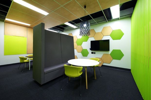 A contemporary commercial collaboration or breakout space with a round white table, lime-green chairs, dark upholstered booth seating, and a wall-mounted television. The room features a black carpeted floor, pale timber acoustic ceiling panels, dark partition walls, bright lime-green ribbed wall cladding, and hexagonal green and timber wall panels. A pendant light and recessed linear lighting provide illumination.