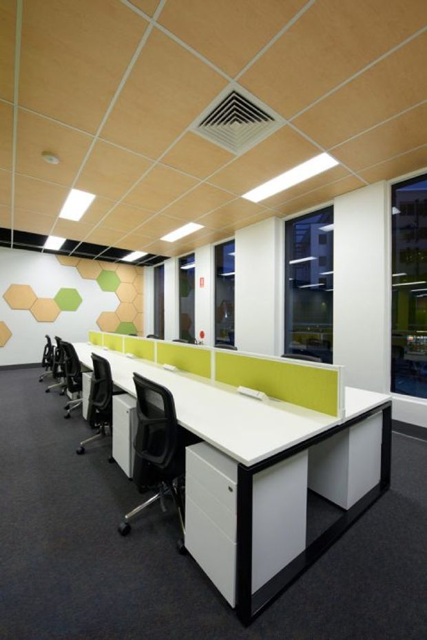 A brightly lit open-plan commercial office with rows of white shared workstations, lime-green privacy screens, black ergonomic office chairs, dark carpet, and tall windows. The ceiling uses tan acoustic panels in a suspended grid with recessed linear fluorescent lighting and a central air-conditioning vent. A rear feature wall has a geometric honeycomb pattern in white, tan, green, and yellow.