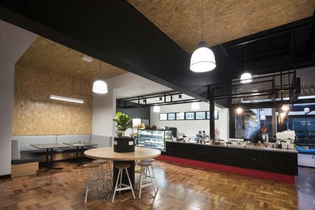 A modern café or commercial hospitality interior with an open service counter, display refrigerator, coffee-making area, communal round tables, banquette seating, and industrial-style black-framed glazing and partitions. The space combines dark structural elements with warm OSB wood-fibre ceiling and wall panels, patterned timber parquet flooring, and bright pendant and linear lighting.