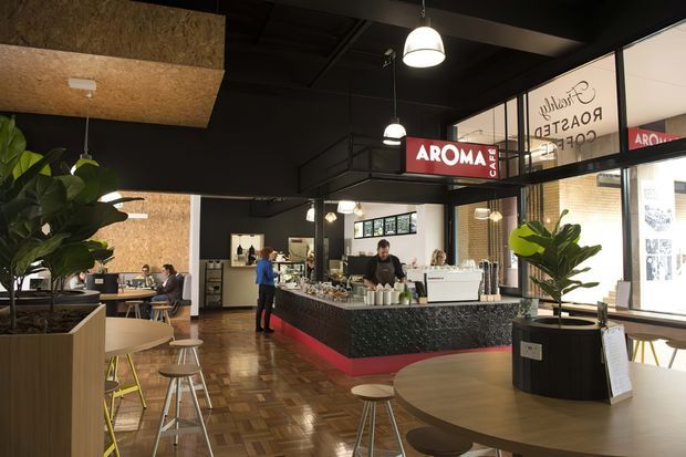 A contemporary café interior with a central service counter, barista station, pendant lighting, timber parquet flooring, black ceiling and structural framing, and large glazed storefront windows. The café features indoor plants, timber tables, stools, and a dark textured counter front with red branding accents.