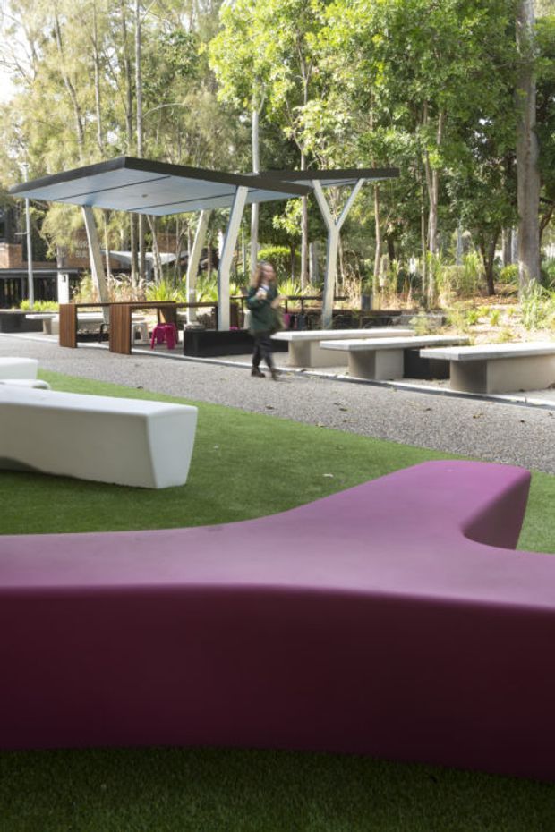 A landscaped outdoor communal space set among mature trees, featuring a large flat-roofed canopy with angled supports, built-in concrete benches, dark timber seating structures, gravel paths, artificial grass, and sculptural white and magenta outdoor furniture. The setting appears to be a designed courtyard or public outdoor amenity area with a contemporary architectural character.