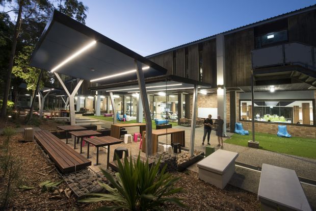 Dusk view of a contemporary multi-level building and landscaped communal outdoor area. The scene features a large covered patio with slim steel posts, integrated linear lighting, timber picnic tables and bench seating, paved paths, and planted garden beds. Glazed ground-floor openings reveal brightly lit interior spaces, while the upper facade uses dark vertical timber cladding and corrugated roofing. The setting appears institutional or commercial rather than residential.