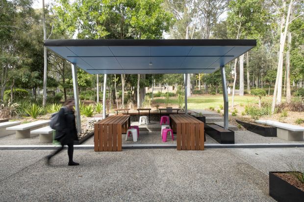 A contemporary outdoor communal dining and seating pavilion set within a landscaped wooded courtyard. The structure has a thin flat canopy roof with dark edging, slender metal posts, built-in concrete bench seating, and multiple timber dining tables and benches. Bright pink stools provide accents beneath the covered shelter, while gravel paving, raised garden beds, and mature trees surround the space.