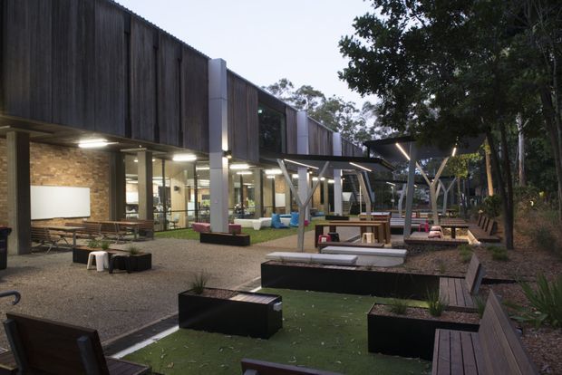 A contemporary multi-level building and landscaped courtyard viewed from outdoors. The facade combines dark vertical timber cladding, exposed brick, large glazed openings, and projecting metal canopy structures. The courtyard contains gravel paving, timber benches and decking, black raised planters, outdoor seating, and a narrow green-surfaced recreational area, surrounded by mature trees.