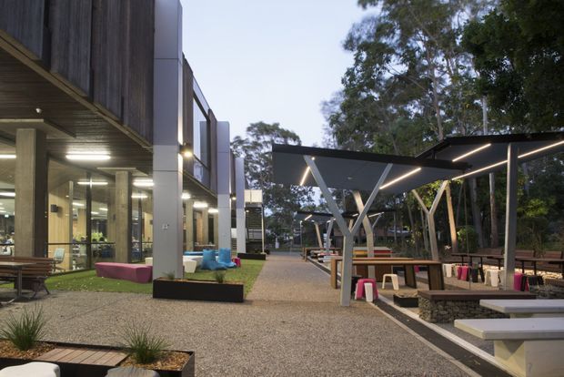 A landscaped outdoor commercial campus or hospitality courtyard viewed at dusk, with a contemporary building on the left and a covered outdoor dining and seating pavilion on the right. The pavilion has a dark flat canopy, exposed angled steel supports, integrated linear lighting, timber tables and benches, and low dark masonry planters. Gravel paving, built-in concrete seating, raised planting beds, and mature trees frame the open space.