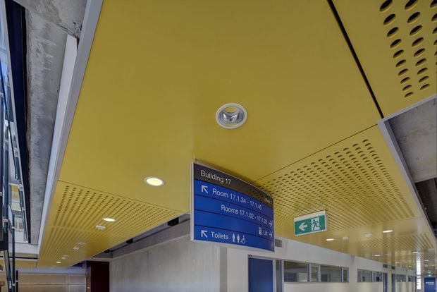 Interior view of a modern institutional or commercial building corridor with a bright yellow perforated metal ceiling, recessed lighting, exposed concrete and glazing, directional blue wayfinding signage, and emergency exit signage. The image focuses on the circulation space and architectural ceiling treatment.