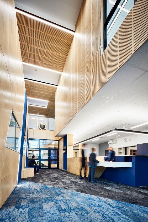 A contemporary commercial reception and lobby interior with a tall atrium-like volume. Light timber slatted wall and ceiling panels, blue-framed glazing and doors, a blue-and-white reception desk, grey and blue patterned carpet, and dark blue accent columns create a bright institutional or corporate entry space. Several people are present around the reception counter, with an upper-level balustrade and clerestory glazing visible above.