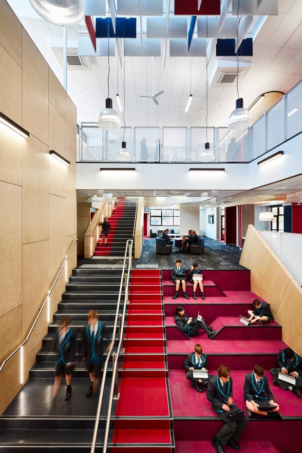 A bright, contemporary multi-level commercial or institutional interior, likely a school or learning facility. The image looks across a large stairway and stepped informal seating area, with students using the vivid magenta and red carpeted tiers for study and socialising. A mezzanine with translucent glass balustrades overlooks the space. The architecture features extensive pale timber wall panelling, white ceilings, exposed hanging pendant lights, linear wall lighting, blue and red suspended acoustic ceiling panels, and a bold red, white, blue and magenta colour scheme.