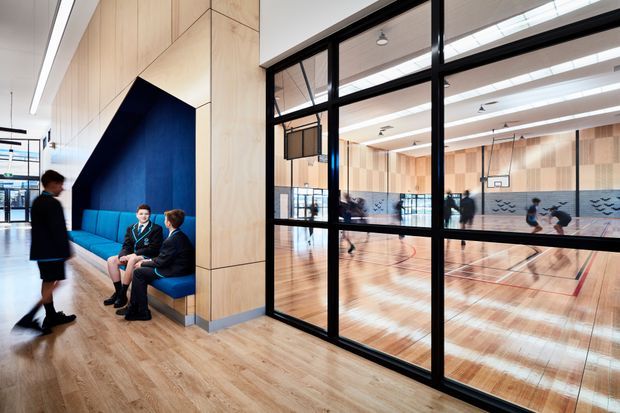 A bright contemporary school or institutional corridor with warm timber flooring and extensive pale timber wall panelling. A built-in blue upholstered bench sits within a dark blue geometric alcove, where several students are seated. Black-framed glazed partitions reveal a full indoor basketball court beyond, with high ceilings, linear lighting, and pale timber acoustic wall finishes.