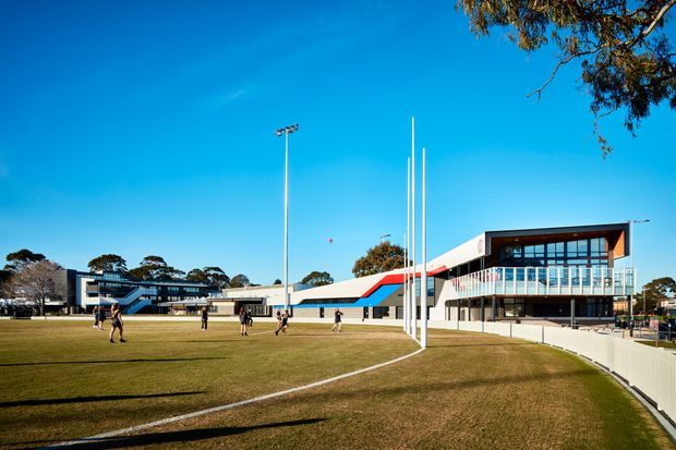 Wide exterior view of a contemporary multi-building sports or educational campus. The low-rise complex has crisp white rendered facades, dark glazed volumes, bold red and blue diagonal facade accents, tall flagpoles, floodlighting, and a large grass sports field in the foreground. People are using the field, while mature trees and a clear blue sky surround the site.