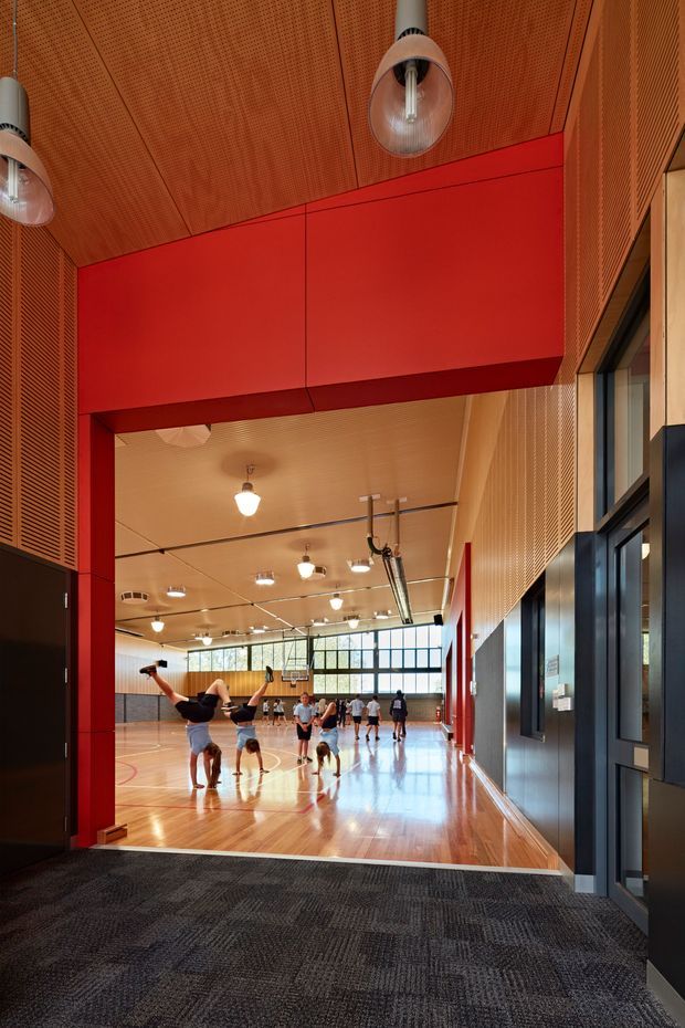 Interior view from a carpeted circulation area into a large indoor basketball court or multipurpose sports hall. The space features a polished timber sports floor, bold red structural framing and wall panels, extensive black-framed glazing, perforated timber acoustic wall and ceiling panels, and suspended industrial-style lighting. People are playing basketball in the background.