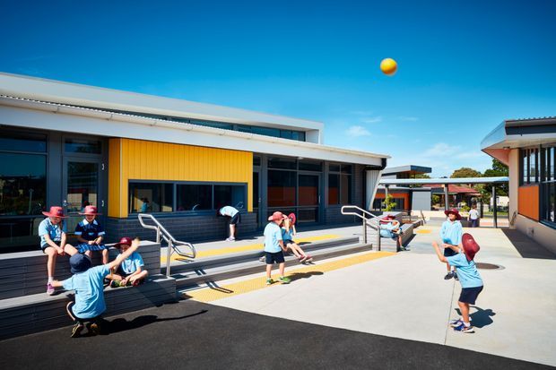 A bright contemporary school or community campus courtyard with low-rise buildings arranged around a paved outdoor space. The main building has horizontal rooflines, dark-framed glazing, grey cladding, a prominent warm yellow vertical-panel feature wall, broad steps with accessible handrails, and tactile paving. Children are playing with a ball in the courtyard, with additional covered walkways and buildings visible beyond.