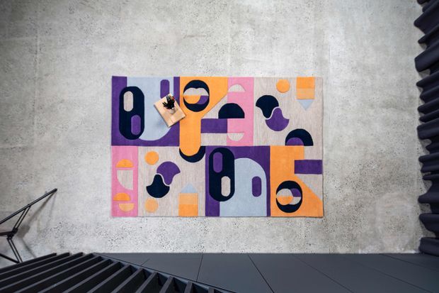 A contemporary abstract geometric artwork mounted on a textured concrete wall, featuring bold blocks and shapes in purple, orange, pink, navy, grey, and cream. The image is viewed from a dark architectural stair or mezzanine edge, with strong linear shadows and industrial-style detailing.