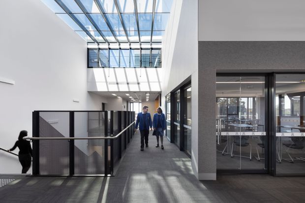 A bright contemporary commercial interior showing a wide circulation corridor beside a stairwell, with black metal balustrades and glazing. A large overhead glass roof or skylight floods the space with daylight and casts linear shadows across the dark floor. On the right, glazed doors reveal a modern meeting or office room with tables and chairs. The architecture is clean-lined, minimal, and institutional or workplace-oriented.