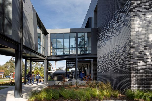 Exterior view of a contemporary multi-storey building arranged around a landscaped pedestrian courtyard. The composition features dark metal framing, extensive glazed connections between wings, and a prominent patterned brick facade in dark grey, light grey, and white. People are visible using the covered walkway, with ornamental grasses and planting in the foreground.