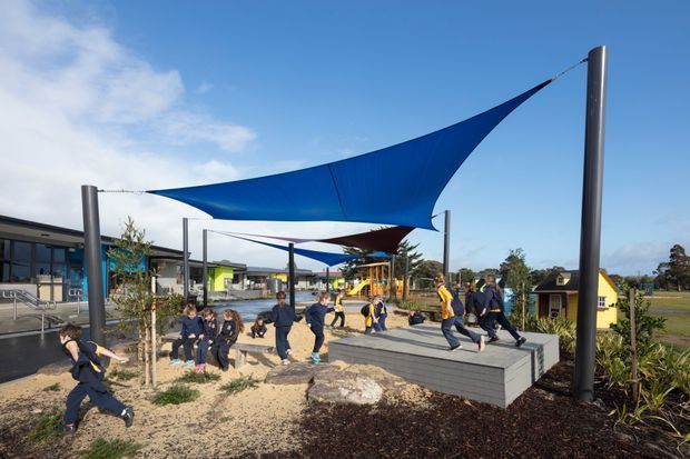 An outdoor recreational area with large blue and burgundy tensioned shade sails supported by tall metal poles. Children are playing and sitting around tiered concrete seating and landscaped planting, with a low modern building and playground structures in the background under a bright blue sky.