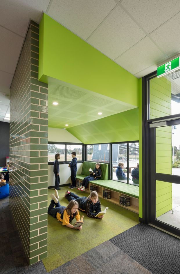 A bright contemporary school or institutional reading and waiting area with vivid lime-green walls, a green glazed brick feature column, built-in upholstered bench seating with open book storage, and children reading or gathering. Large black-framed windows and a glazed entrance provide abundant daylight, while a suspended acoustic tile ceiling and carpeted floor support the public interior fit-out. An emergency exit sign is visible above the doorway.
